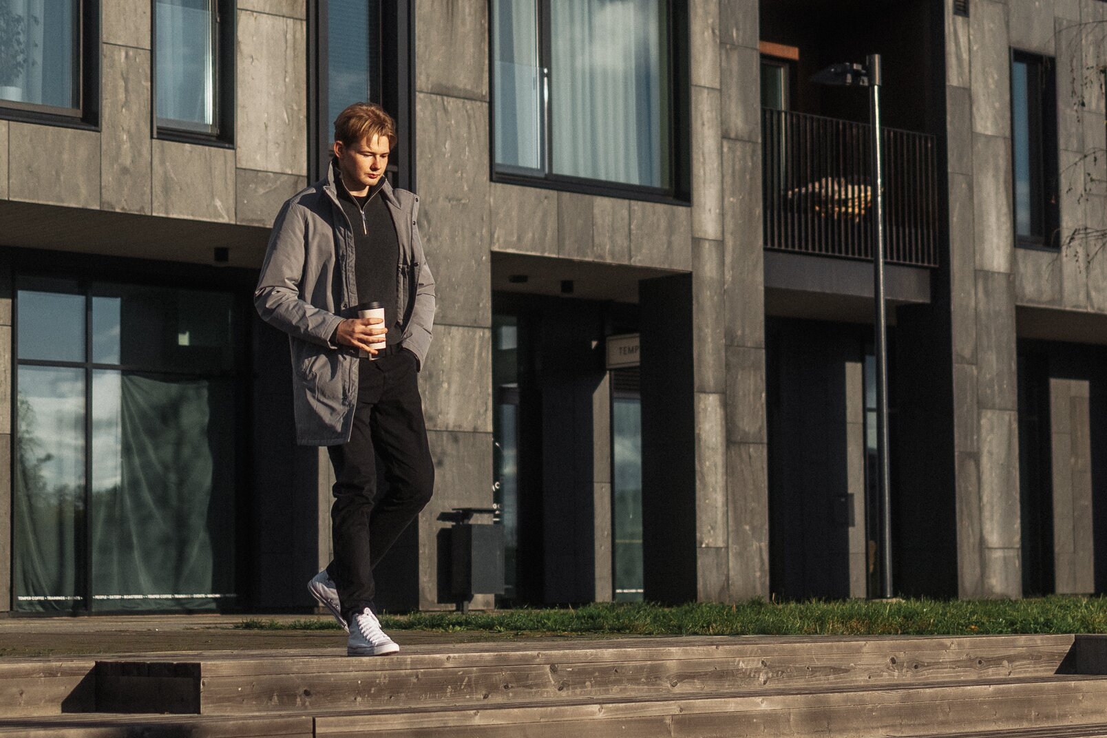 A young man in a gray jacket and black pants holds a coffee cup while walking in front of a modern building with large windows and a balcony, illuminated by natural light.